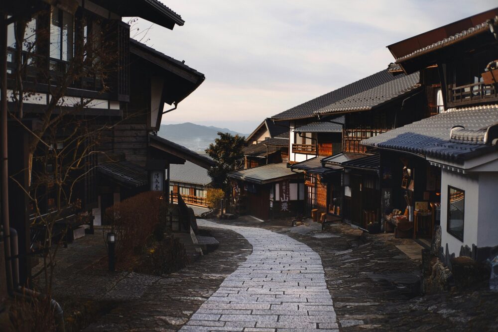 Scenic view of a traditional Japanese village street with wooden houses and tiled roofs, surrounded by a peaceful mountain backdrop.