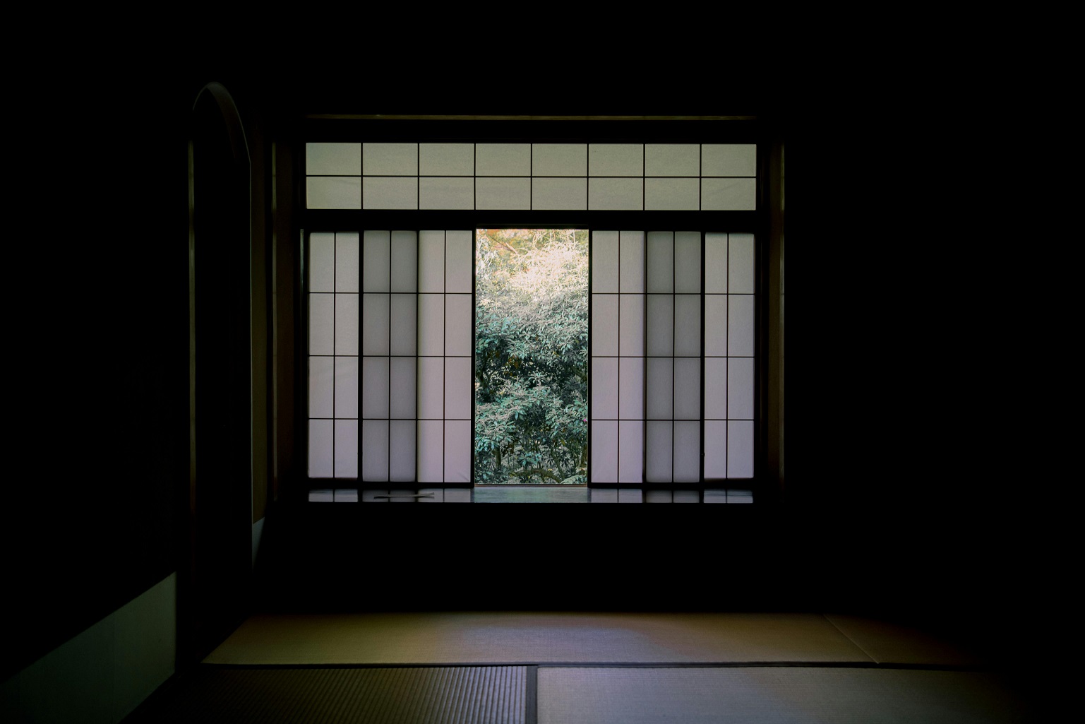 Minimalist Japanese room with tatami flooring and a shoji window framing a serene view of greenery.