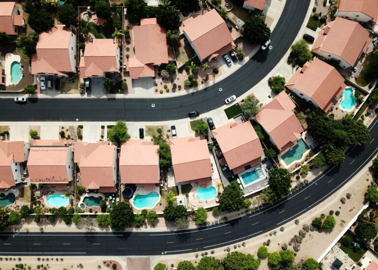 Top-down view of a suburban neighborhood featuring houses with red roofs, driveways, and backyard swimming pools arranged along curved roads.