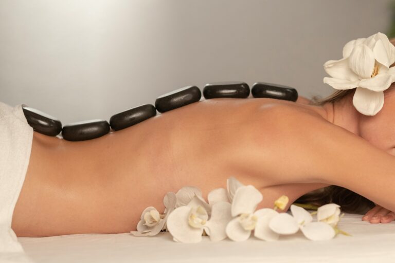 Woman receiving a hot stone massage therapy with black stones aligned along her back, surrounded by white orchids.